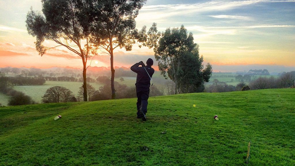 A person swings a golf club on a grassy course at sunrise or sunset, with trees and a picturesque landscape in the background—an inspiring moment reminiscent of charity tournaments supporting Galway’s Sick Kids.