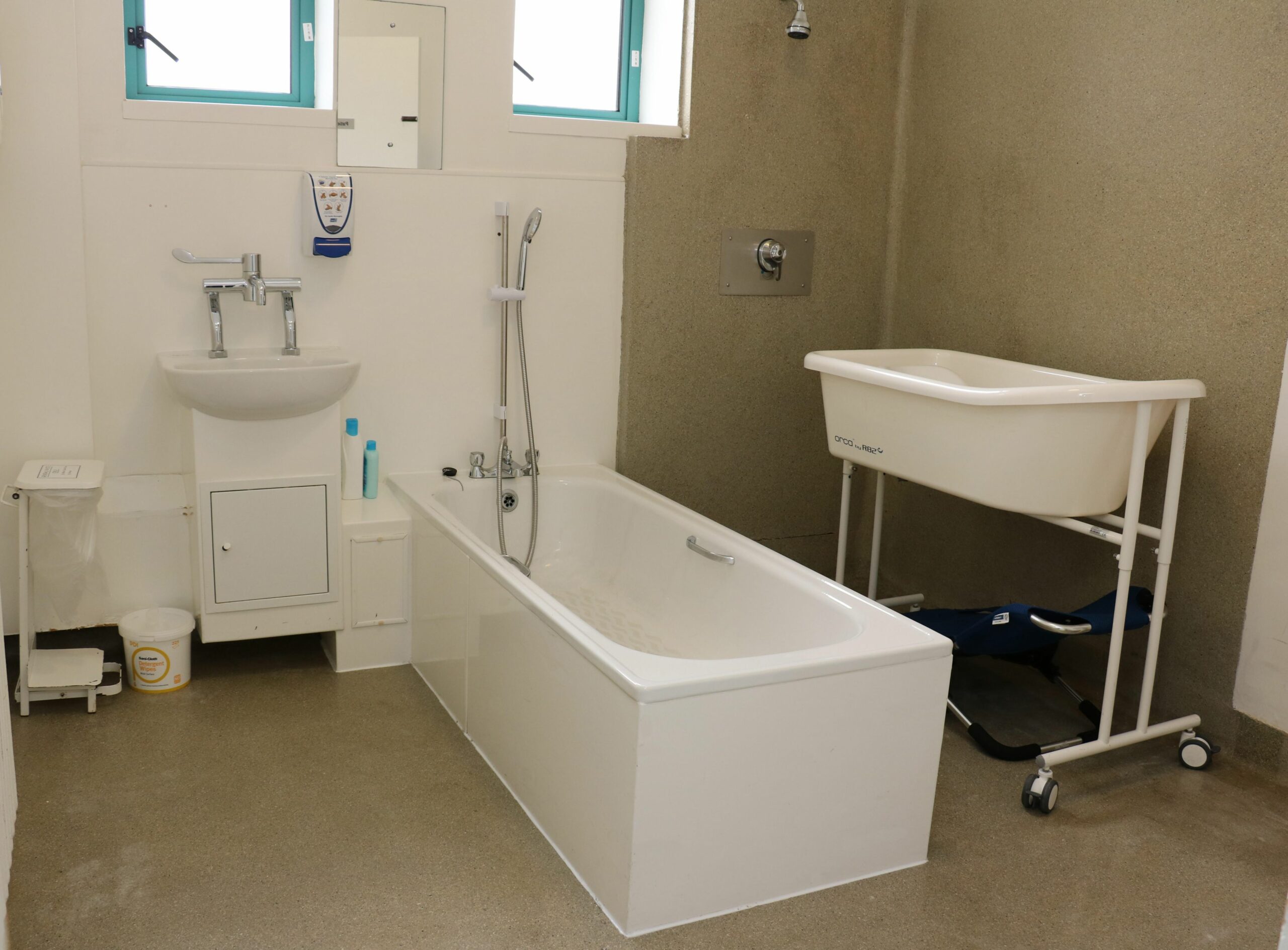 A clean bathroom with a white bath, basin, and a baby changing table on wheels, thoughtfully set up for Galways Sick Kids. There are two bottles on the floor and a towel rail on the wall near a window.