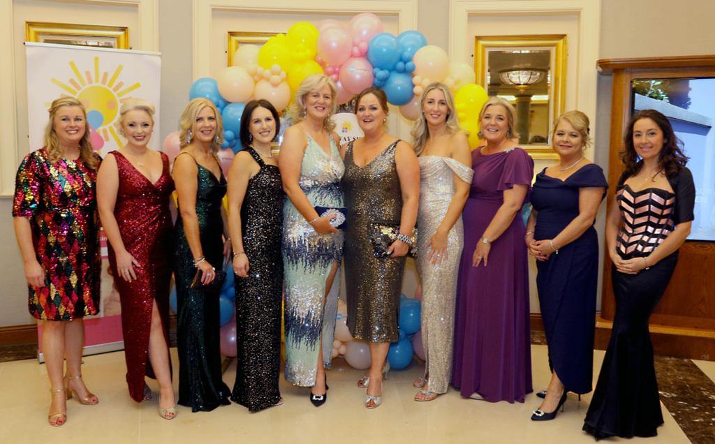Ten women in formal evening dresses stand together, smiling in front of colourful balloons and a banner with a sun design, at an indoor event supporting Galway Sick Kids.