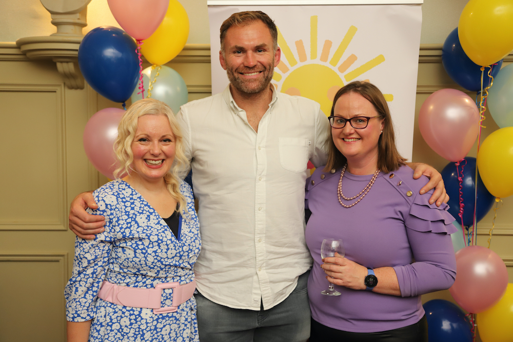 Three adults stand smiling at a cheerful indoor event with colourful balloons and a sun poster in the background. One person holds a drink, and all appear happy and relaxed as they gather in support of Galways Sick Kids.
