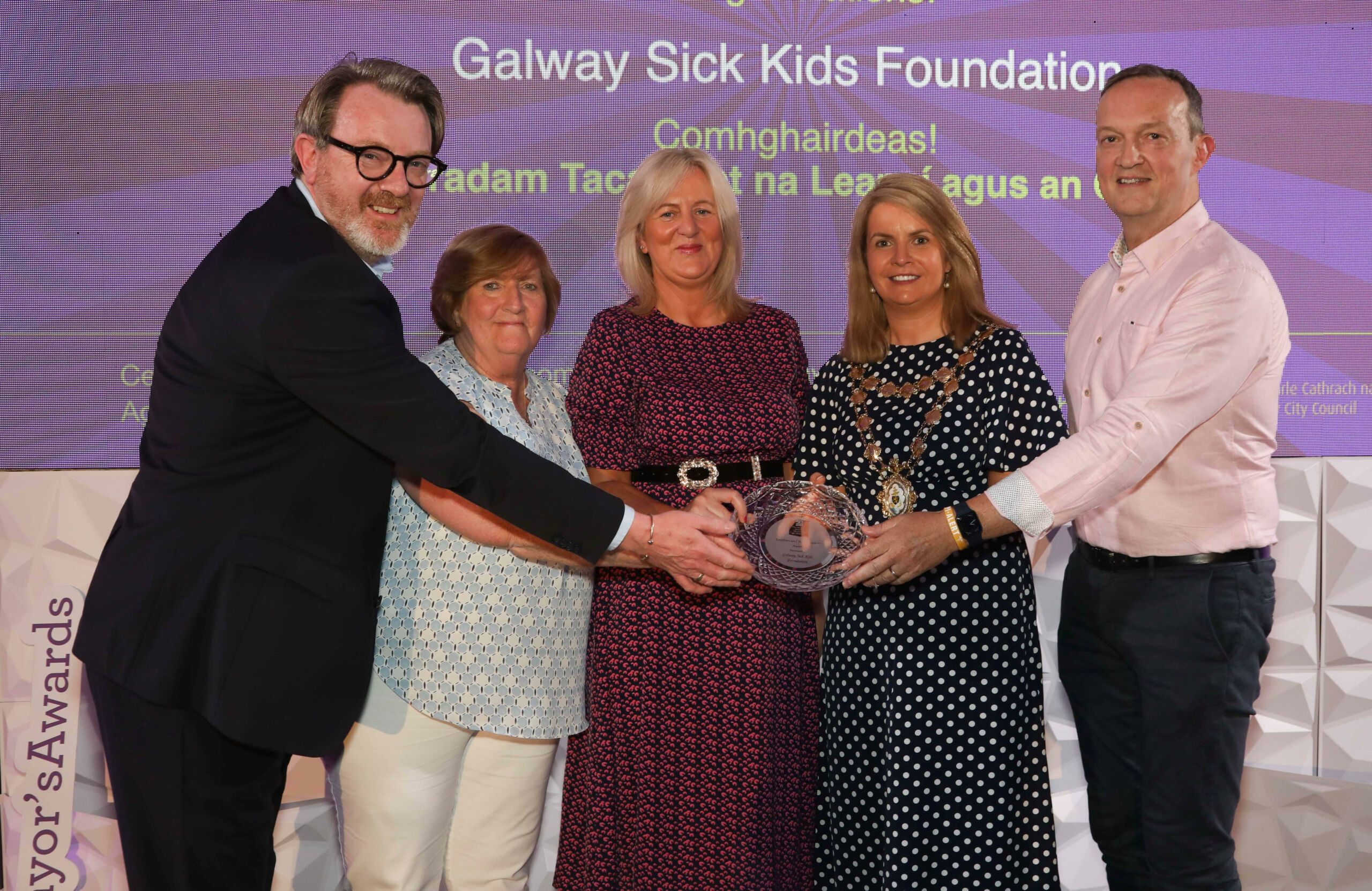Five people stand together on stage, smiling and holding a glass award. A background screen displays Galway’s Sick Kids Foundation and other text. The group, representing Galway’s Sick Kids, is dressed in business casual attire at an awards event.