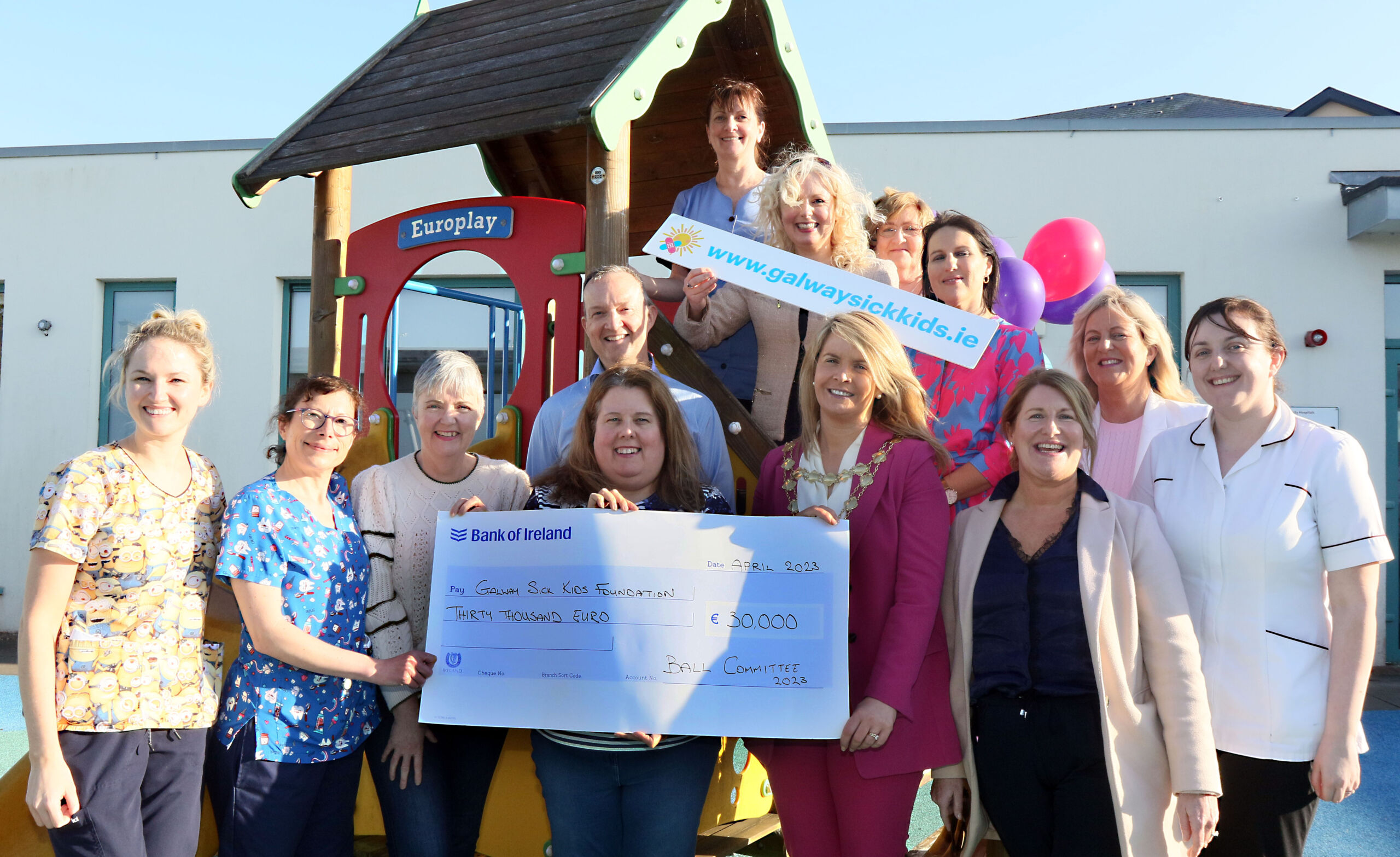 A group of smiling adults pose outside by a playhouse, holding a large cheque for €10,000 and a colourful www.galwaysickkids.ie sign. Some wear medical uniforms, celebrating a generous donation in support of Galway Sick Kids.