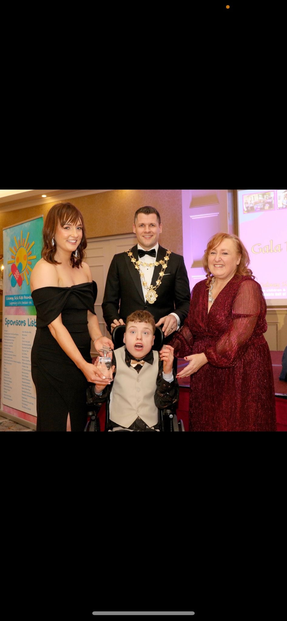 Four people, including one in a wheelchair, pose at an awards event celebrating achievements related to Galway’s Sick Kids. Two women hold glass awards, a man in a chain of office stands behind them, and a sponsor list is visible in the background.