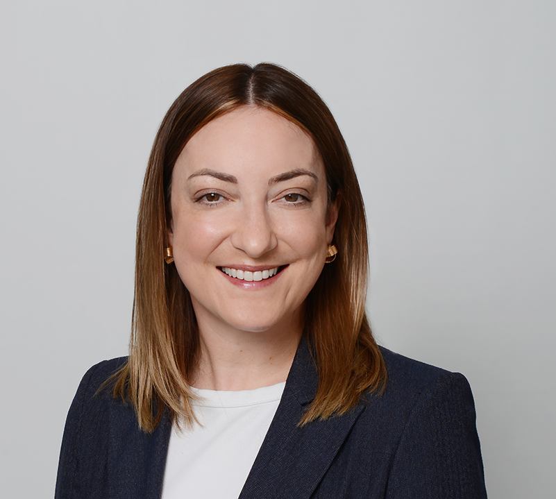 Smiling woman with straight, shoulder-length brown hair, wearing a navy blazer and white top, posed against a plain light grey background—her warm expression echoes the compassion often seen in supporters of Galway’s Sick Kids.