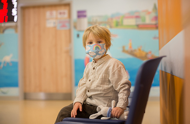 A young child wearing a face mask sits on a chair in a brightly coloured room, holding a soft toy, likely in a hospital or clinic waiting area, embodying the resilience of Galway's Sick Kids.