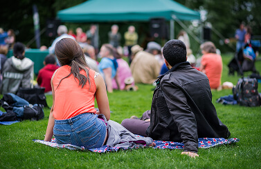 Two people sit on a blanket on grass, facing a blurred outdoor event with more people and a green marquee in the background, possibly attending a gathering in support of Galway’s Sick Kids.