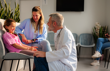 A doctor kneels and smiles whilst talking to a young girl and her mother in a waiting room at Galway’s Sick Kids. The mother and daughter are seated, and plants decorate the area.