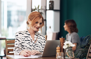 Woman with red hair sitting at a table, working on a laptop and writing on paper. She is smiling, perhaps working on an initiative for Galway’s Sick Kids, with a blurred person in the background and a vase of flowers on the table.