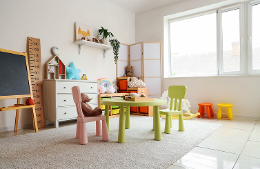 Bright playroom with a green table, pastel chairs, toys, soft toys, shelves, and a large window letting in natural light. Designed to bring comfort and cheer for Galways Sick Kids, a soft rug covers part of the floor.