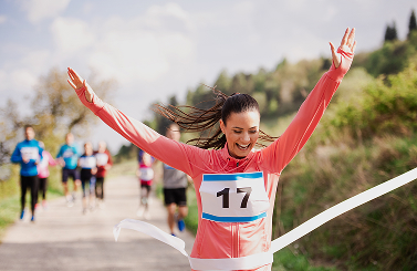 A woman wearing a race bib number 17 raises her arms in victory as she crosses the finish line outdoors, proudly supporting Galways Sick Kids, with other runners in the background.