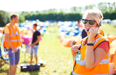 A smiling person in an orange safety vest and headset stands outdoors at a Galways Sick Kids event, with blurred people and marquees in the background under bright sunlight.