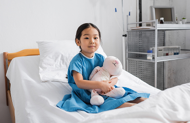 A young girl in a hospital gown sits on a bed, holding a white soft toy. Medical equipment is visible in the background, illustrating the quiet resilience often seen among Galway’s Sick Kids.