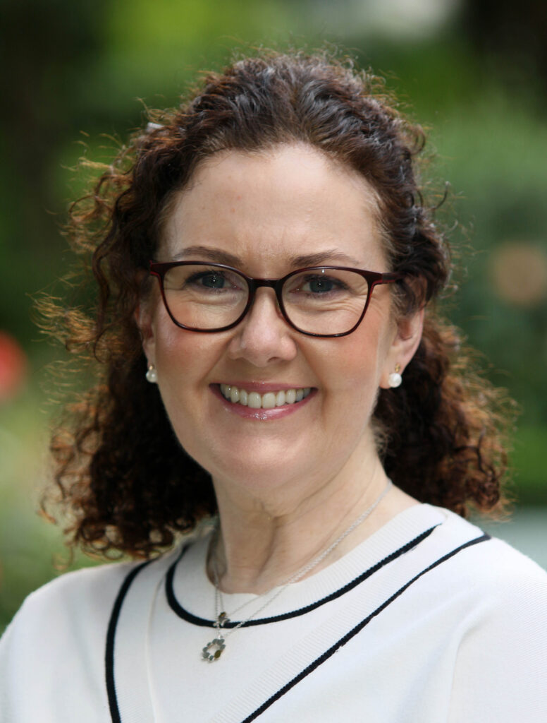 A woman with curly brown hair, glasses, and a white top smiles outdoors, the lush greenery softly blurred in the background—a warm moment captured during her voluntary work with Galway’s Sick Kids.