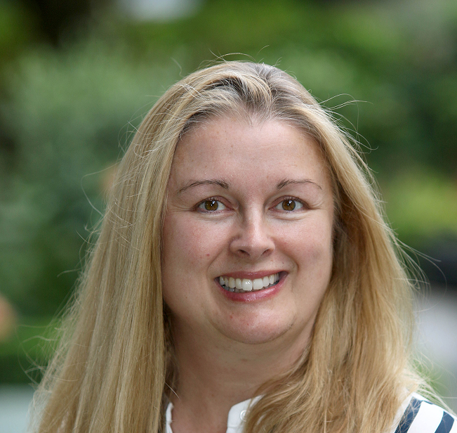 A woman with long blonde hair smiles at the camera, radiating warmth and positivity. She is outdoors with greenery blurred in the background, embodying the spirit of support for Galway’s Sick Kids.