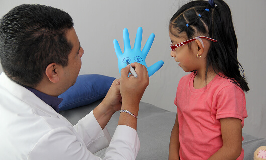 A doctor from Galway’s Sick Kids draws a smiley face on a blue glove for a young girl with glasses, who is watching and smiling.