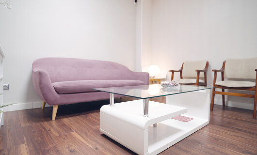 A modern waiting room designed to welcome Galway's Sick Kids features a pink sofa, two wooden chairs with beige cushions, a glass coffee table, and a lamp, all set on a wooden floor against a plain white wall.