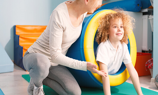 Child crawling through a large, yellow and blue foam ring while an adult from Galways Sick Kids guides them, both indoors on a padded floor with play equipment in the background.