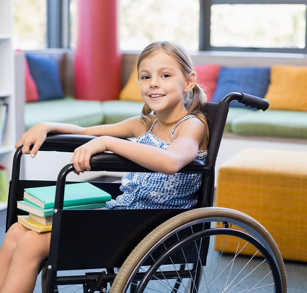 Smiling young girl in a wheelchair holds books on her lap, sitting in a bright room with colourful cushions and a window in the background, representing the cheerful spirit of Galways Sick Kids.