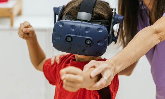 A child wearing a VR headset raises their arms while an adult, partially visible, helps guide their movements. The child, dressed in a red shirt, is one of Galway's Sick Kids engaging in this innovative, supportive activity.
