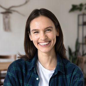 Young woman with straight brown hair smiling, wearing a blue checked shirt over a white t-shirt, sitting indoors with blurred plants and shelves in the background—her warmth and positivity reflect the supportive spirit of Galways Sick Kids.