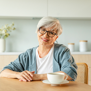 Smiling older woman with short grey hair and glasses sits at a table with a coffee cup in front of her, in a bright, modern kitchen, as she reflects on her recent fundraising efforts for Galways Sick Kids.