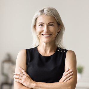 Smiling woman with fair hair in a sleeveless black top stands with her arms folded, facing the camera, in a softly lit room with a blurred background—capturing the caring spirit behind Galways Sick Kids.