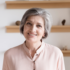 Smiling older woman with short grey hair wearing a light pink blouse stands indoors in front of wooden shelves with small decorative objects, showing her support for Galways Sick Kids.