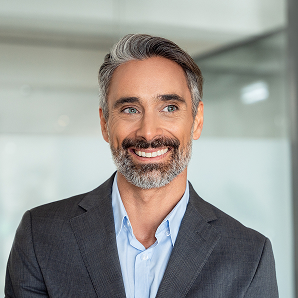 Smiling man with grey hair and beard wearing a grey suit jacket and light blue shirt, standing indoors in a modern office setting with glass walls in the background, shows his support for Galways Sick Kids.
