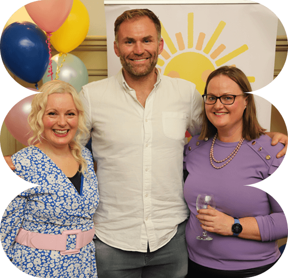 Three adults smile at a gathering in support of Galways Sick Kids, standing in front of a colourful balloon display and a sun-themed banner. One woman holds a drink, while another wears a blue patterned dress. The man in the centre wears a white shirt.