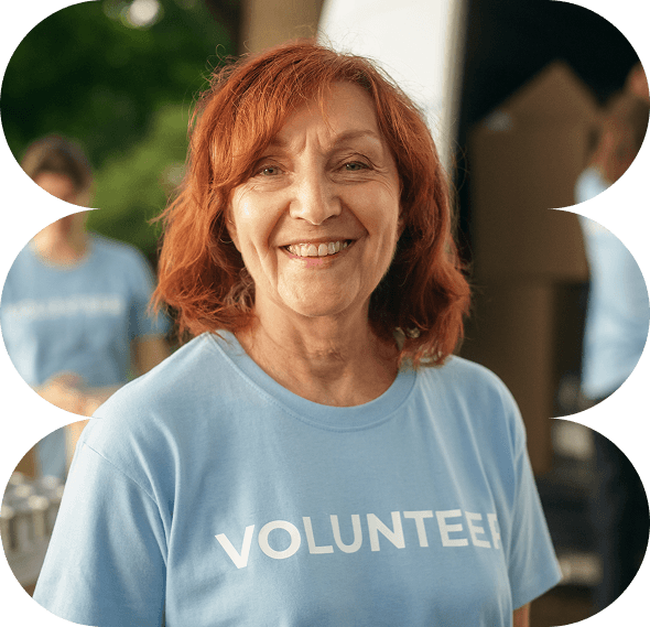 Smiling woman with red hair wearing a blue VOLUNTEER shirt stands outdoors, supporting Galway's Sick Kids, with other volunteers blurred in the background.