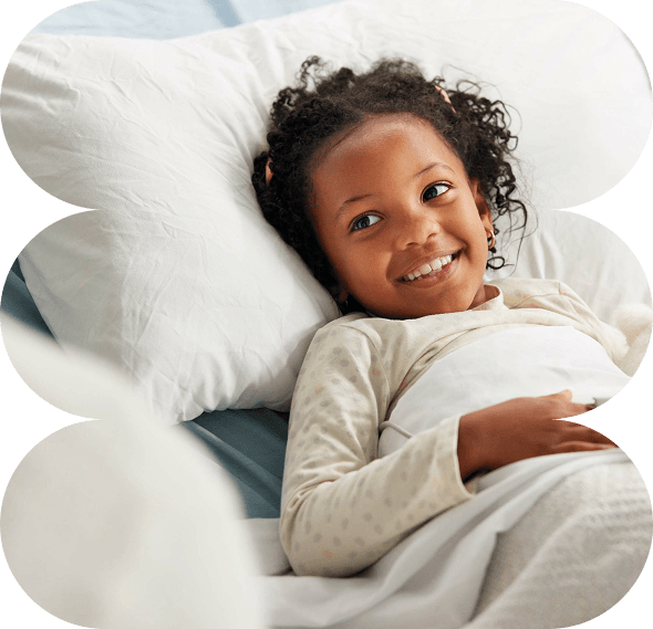 Smiling young girl with curly hair lies in bed under a white duvet, resting her head on a pillow and looking slightly upward—capturing the hopeful spirit of Galway's Sick Kids.