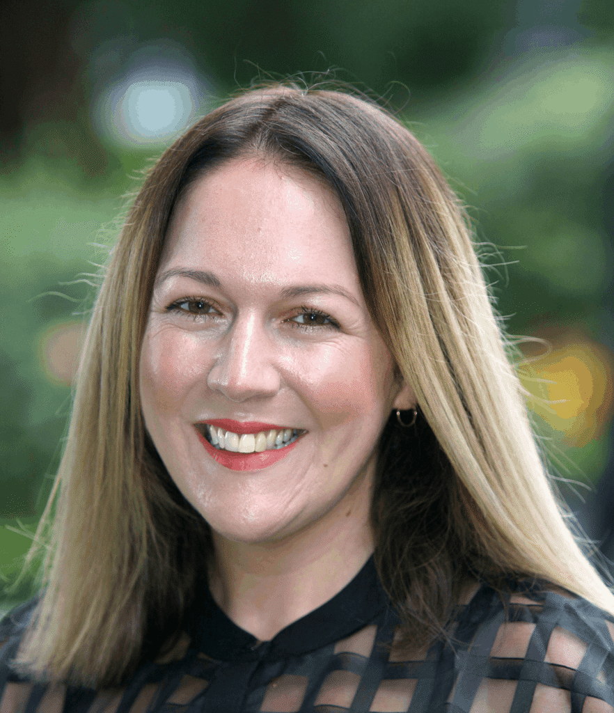 A woman with straight, shoulder-length blonde hair smiles outdoors, wearing a black top with a chequered pattern. The background is blurred greenery, giving the scene a warm and uplifting atmosphere reminiscent of Galway’s Sick Kids’ inspiring community spirit.
