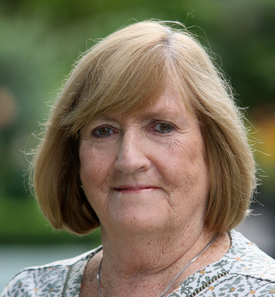 An older woman with short, light brown hair and a patterned blouse smiles gently at the camera. The background is blurred greenery, adding a warm, hopeful touch that echoes the compassionate spirit of Galway's Sick Kids.